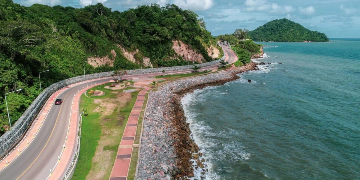 Coastal road along Thailand eastern seaboard with turquoise ocean views, fishing boats, and palm trees lining the route